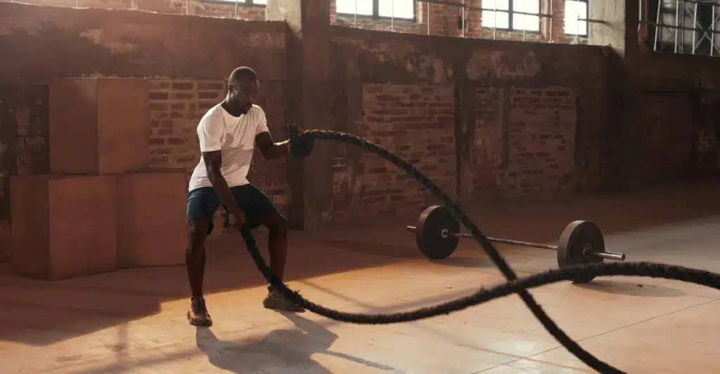 A man in a white t-shirt and blue shorts is vigorously swinging heavy battle ropes in a gym with large windows and a concrete floor.