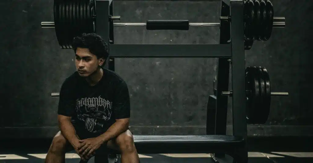 A man in a black t-shirt and shorts is sitting on a gym bench, looking off to the side with a pensive expression, with weight racks visible behind him.