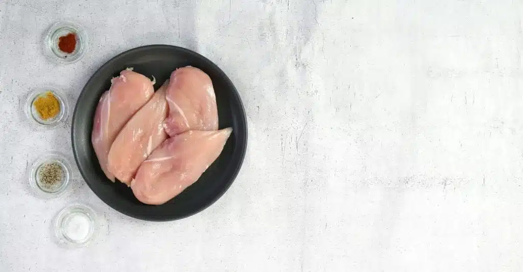 An overhead view of four raw chicken breasts on a black plate, accompanied by small glass containers of various spices including paprika, curry powder, pepper, and salt, all set on a light gray surface.