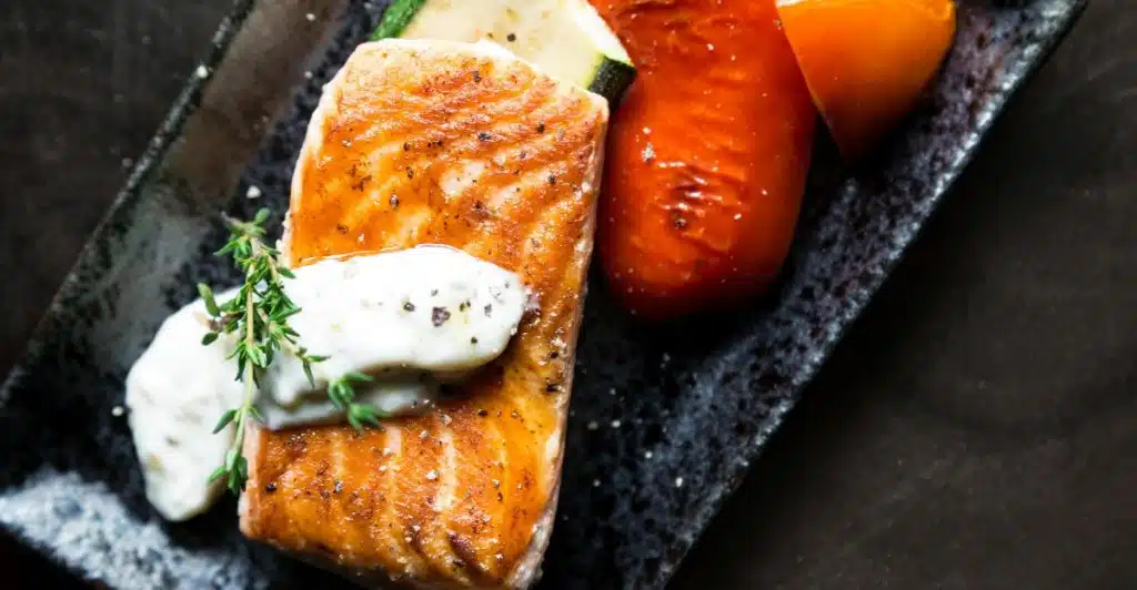 A perfectly grilled salmon fillet, topped with a dollop of white sauce and a sprig of thyme, is presented on a dark textured plate alongside grilled zucchini and roasted red and yellow bell peppers.