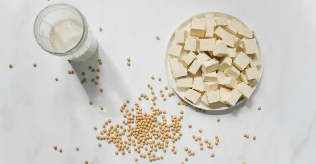 An aerial shot shows a white plate filled with neatly sliced cubes of tofu next to a glass of soy milk, with a scattering of soybeans across the light-colored surface.