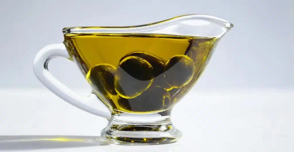 A clear glass gravy boat is filled with golden olive oil, with several dark olives submerged at the bottom, all against a bright background.
