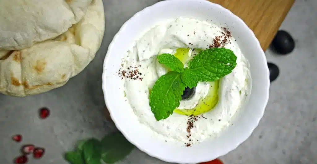 A white bowl holds creamy Greek yogurt garnished with green mint leaves and black olives, sprinkled with a dark spice, set next to flatbread, cherry tomatoes, and pomegranate seeds on a gray surface.