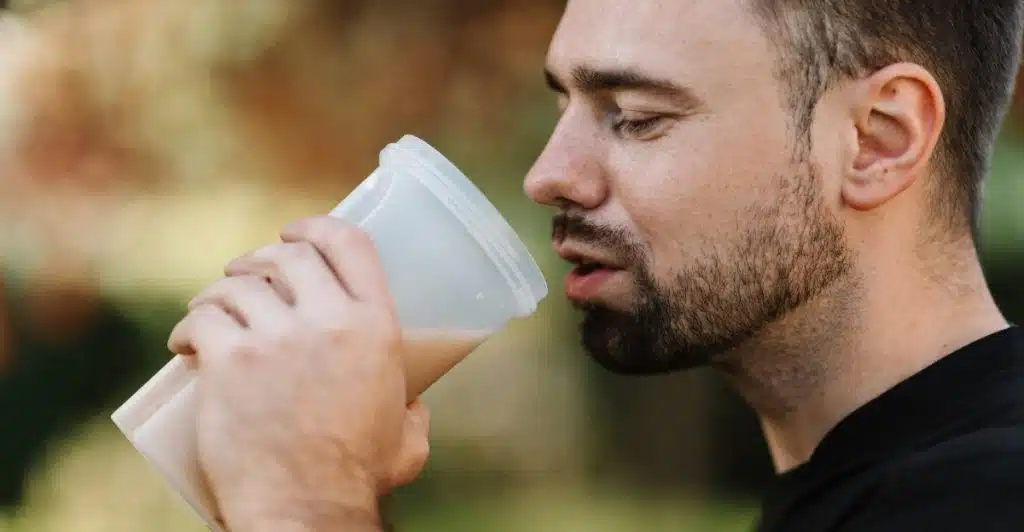 A man with short dark hair and a beard, wearing a black shirt, is shown in profile as he drinks from a clear plastic shaker bottle, with a blurred green background.