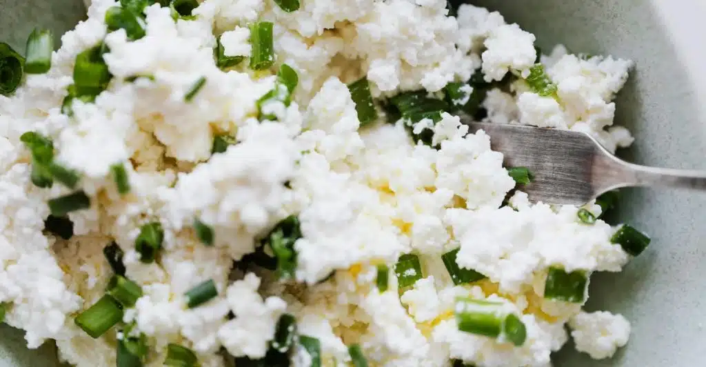 A close-up, high-angle view of a bowl filled with fluffy white cottage cheese, generously mixed with chopped fresh green onions, with a fork partially visible.