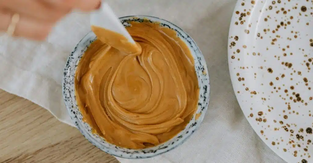 A hand holds a small white spatula, gently swirling creamy peanut butter in a small, patterned bowl, with a speckled white plate and a white linen cloth visible nearby.