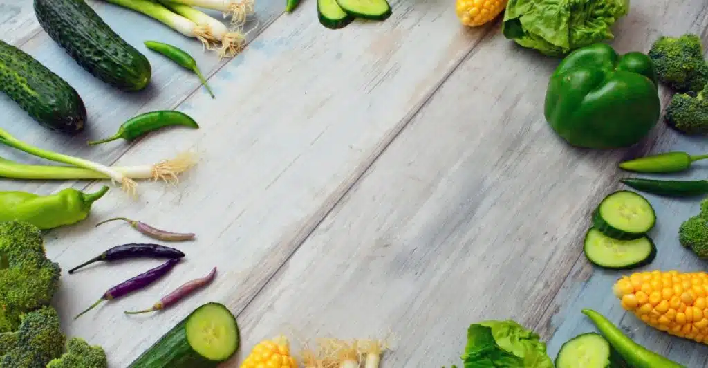 A collection of fresh, colorful vegetables including cucumbers, green onions, various peppers, corn, lettuce, and broccoli are artfully arranged around an empty space on a rustic brown wooden table.
