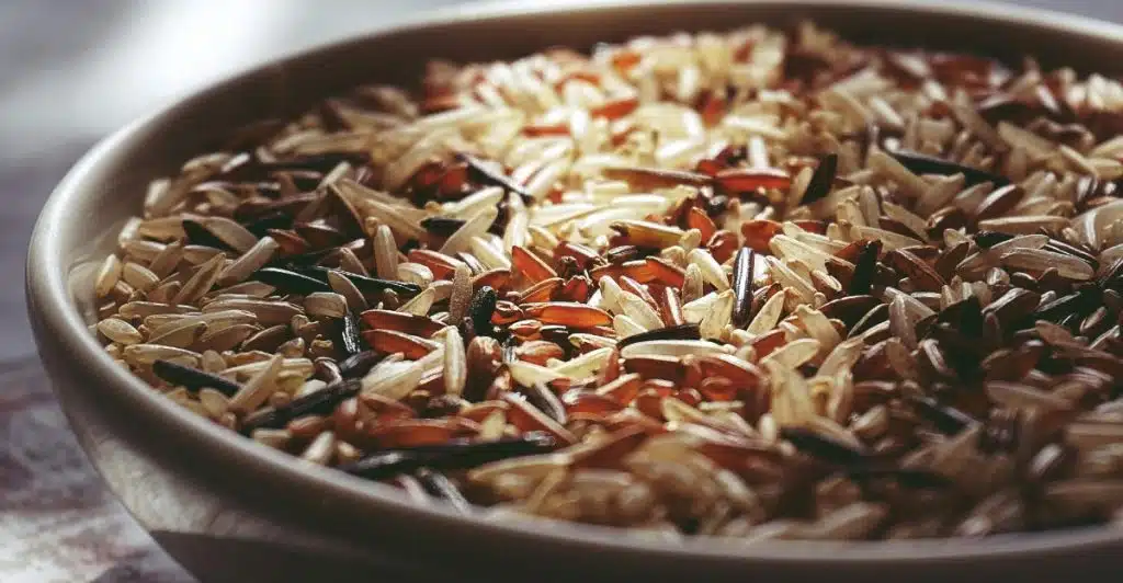 A close-up, slightly angled view of a white bowl filled with a mix of light brown and dark brown rice grains, catching the natural light.