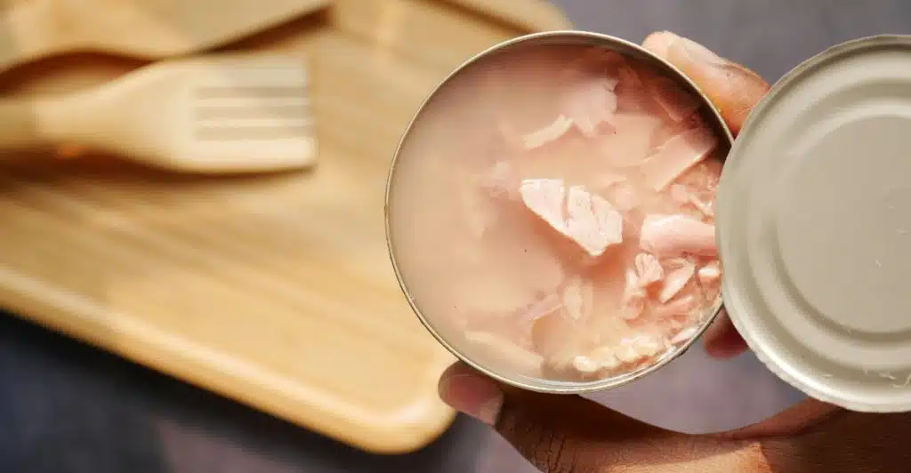 A person's hand holds an open, round tin can filled with flaked light pink tuna meat in water, with a wooden serving board and utensils blurred in the background.