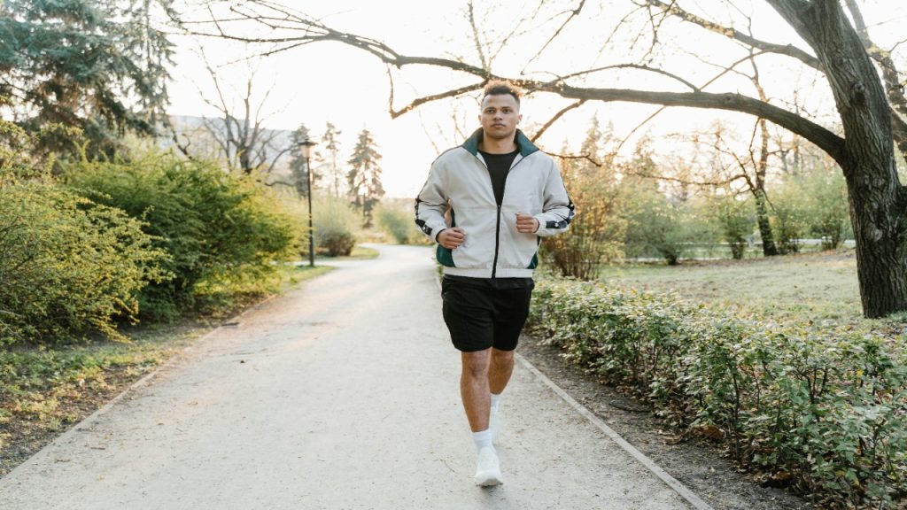 A young man jogs along a tree lined path in a park during early morning.