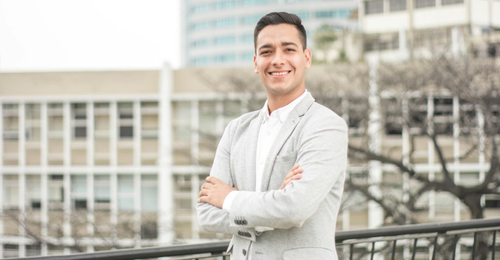 A confident young man stands outdoors with his arms crossed, smiling warmly.