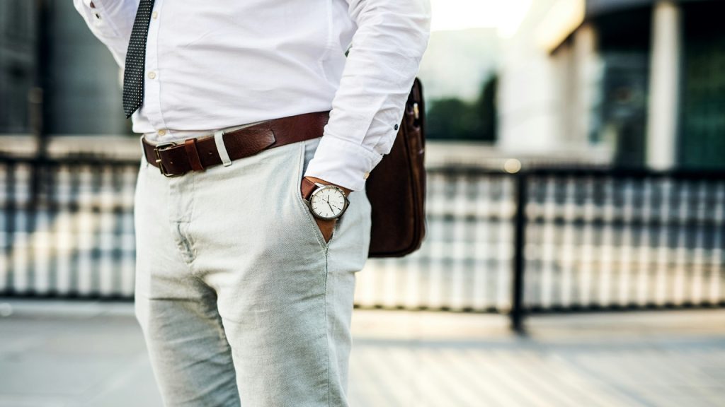 A man wearing a white dress shirt, brown belt, and light trousers with his hand in his pocket.