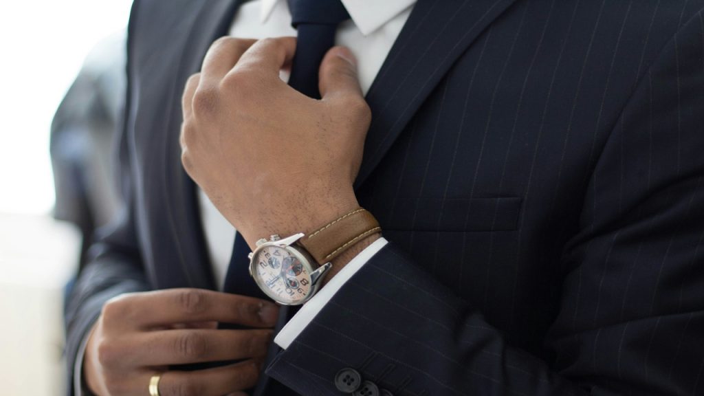 A man adjusts his necktie and wears a black suit, with a brown dress watch on his wrist.