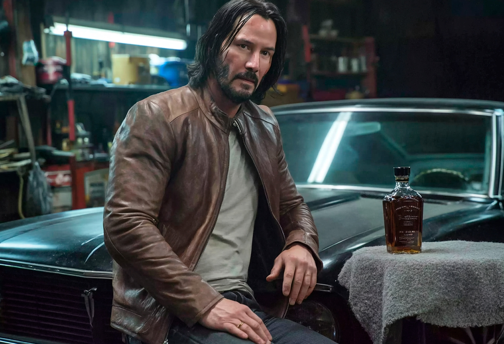 John Wick in brown leather jacket and gray shirt leaning on black car in garage with bottle of whiskey