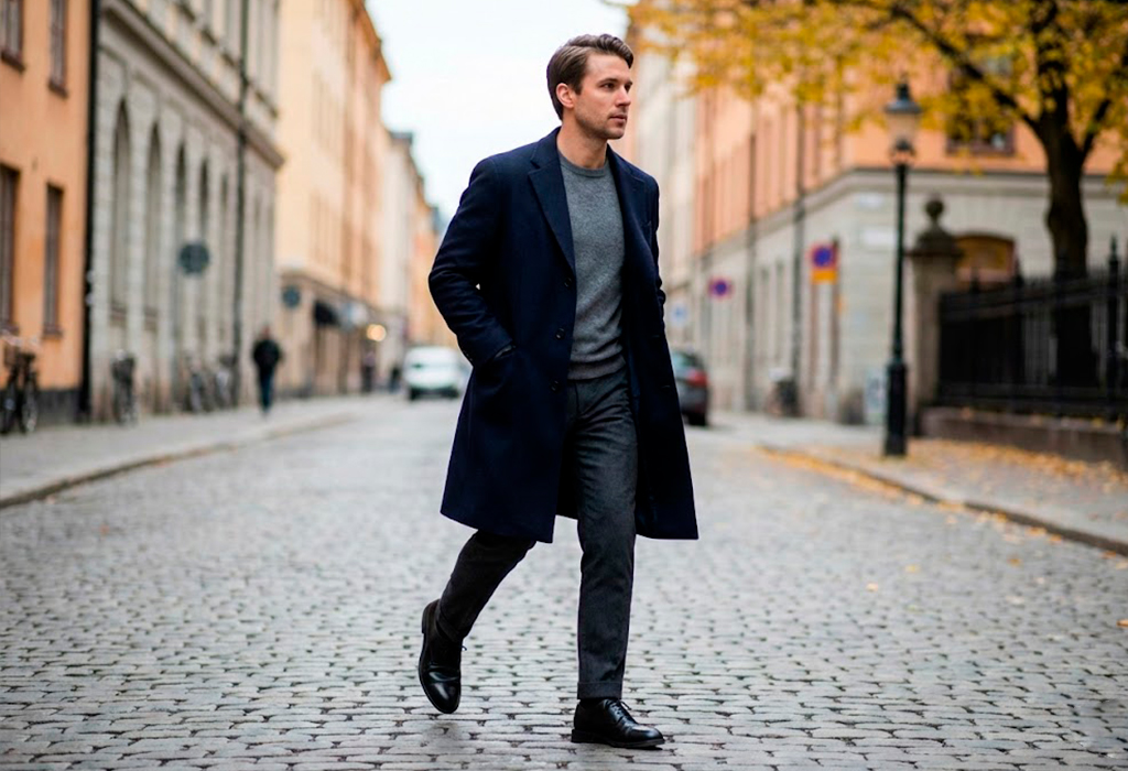 Man wearing a navy overcoat styled with a grey sweater, tailored trousers, and leather shoes in fall.